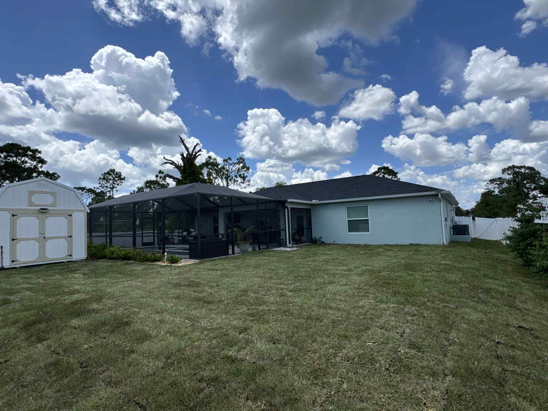 Backyard with a light blue house, screened patio, white shed, and partly cloudy sky.