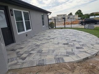 Stone patio with a curved edge next to a gray house with a dark door. A black fence in the background.