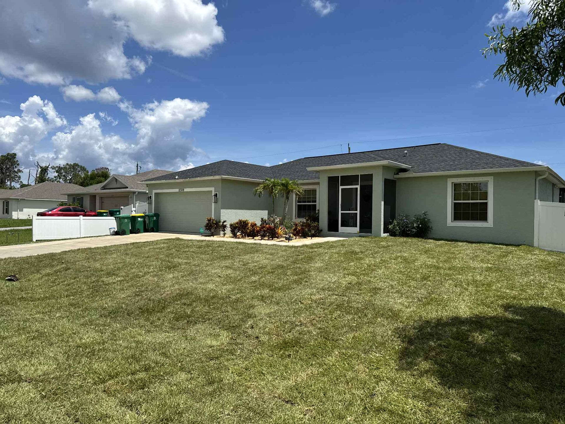 Light green house with lawn, blue sky, clouds. Driveway with green bins.