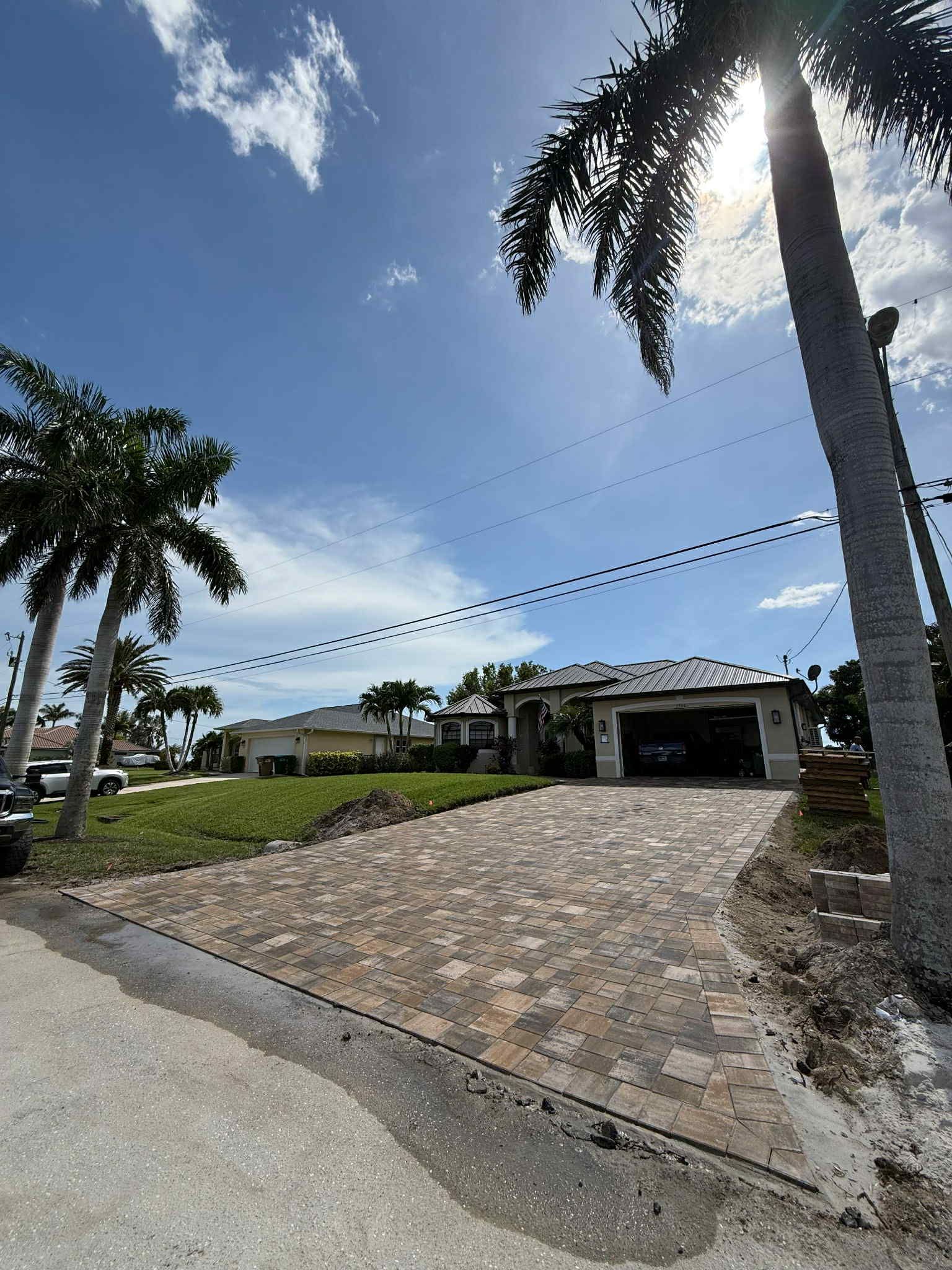 A sunny day in front of a house with a brick driveway, palm trees, and a blue sky.