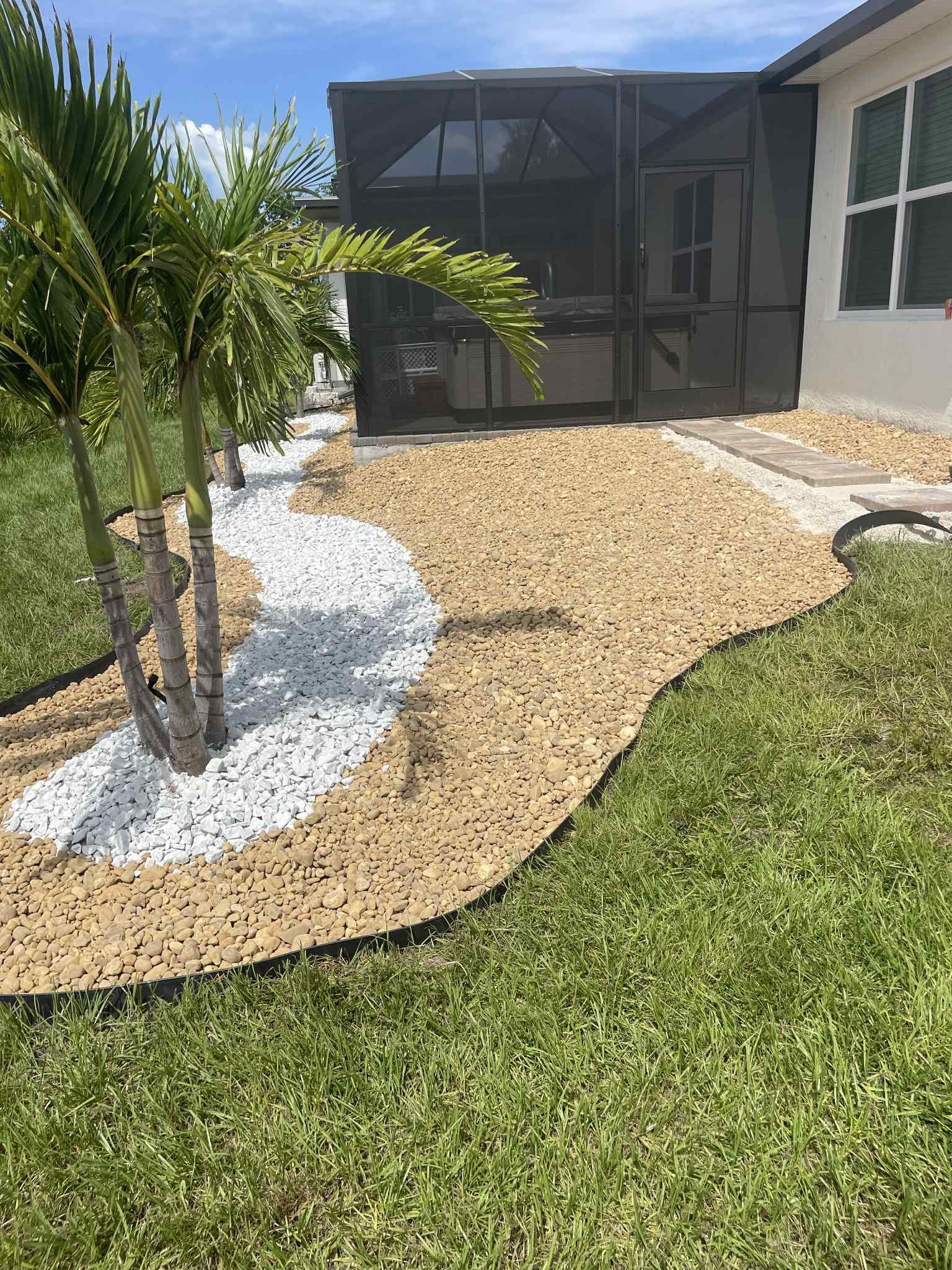 Pathway with tan and white gravel next to a house, palm trees, and a screened-in patio.