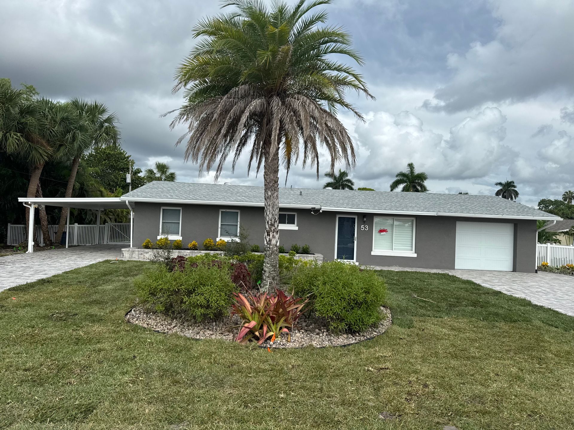 Gray house with carport, white garage door, and palm tree in front yard. Overcast sky.