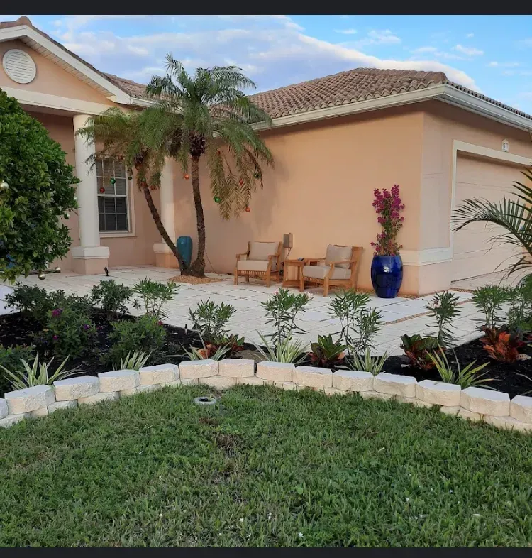 Beige house with palm trees, lawn, and flowerbeds. Outdoor seating, pink flowers in blue pot.