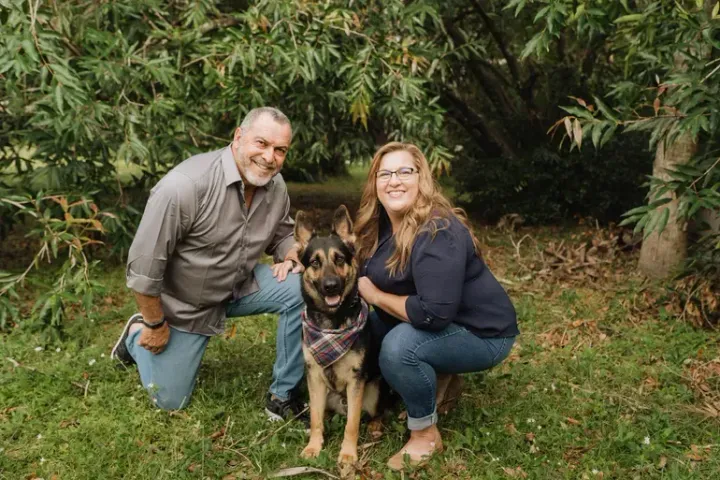 Man and woman kneeling with German Shepherd dog in a grassy outdoor setting; all are smiling.