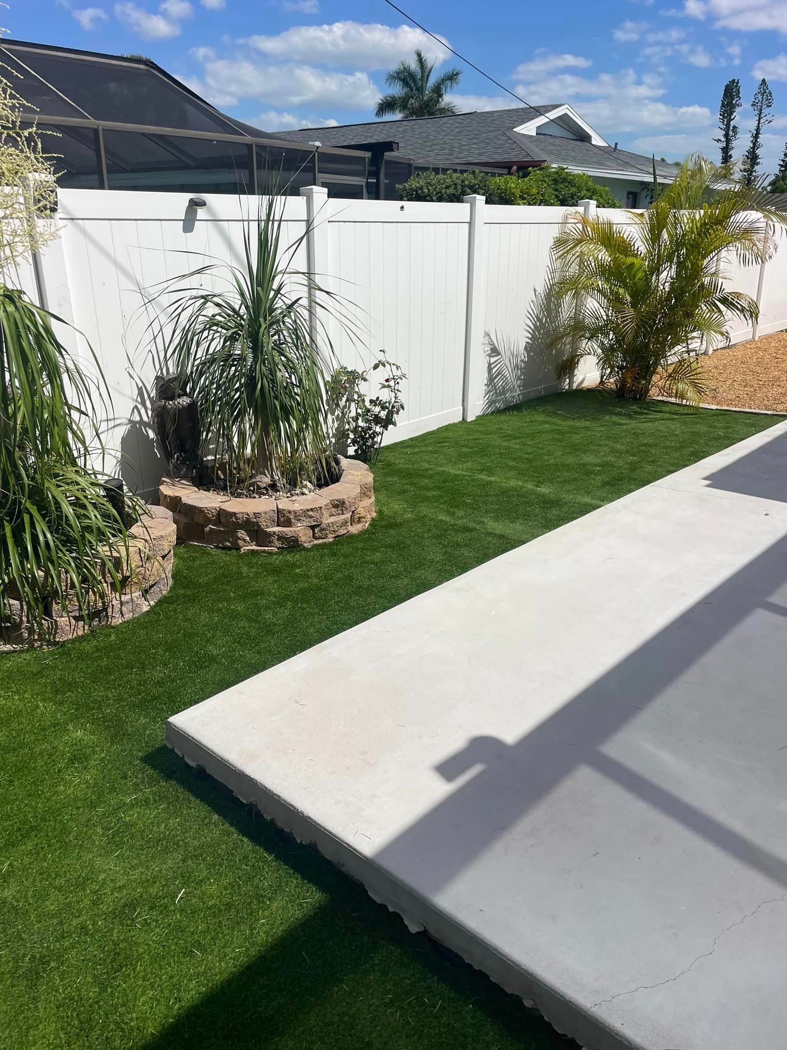 Backyard with white fence, green grass, concrete patio, and plants. Sunny day.