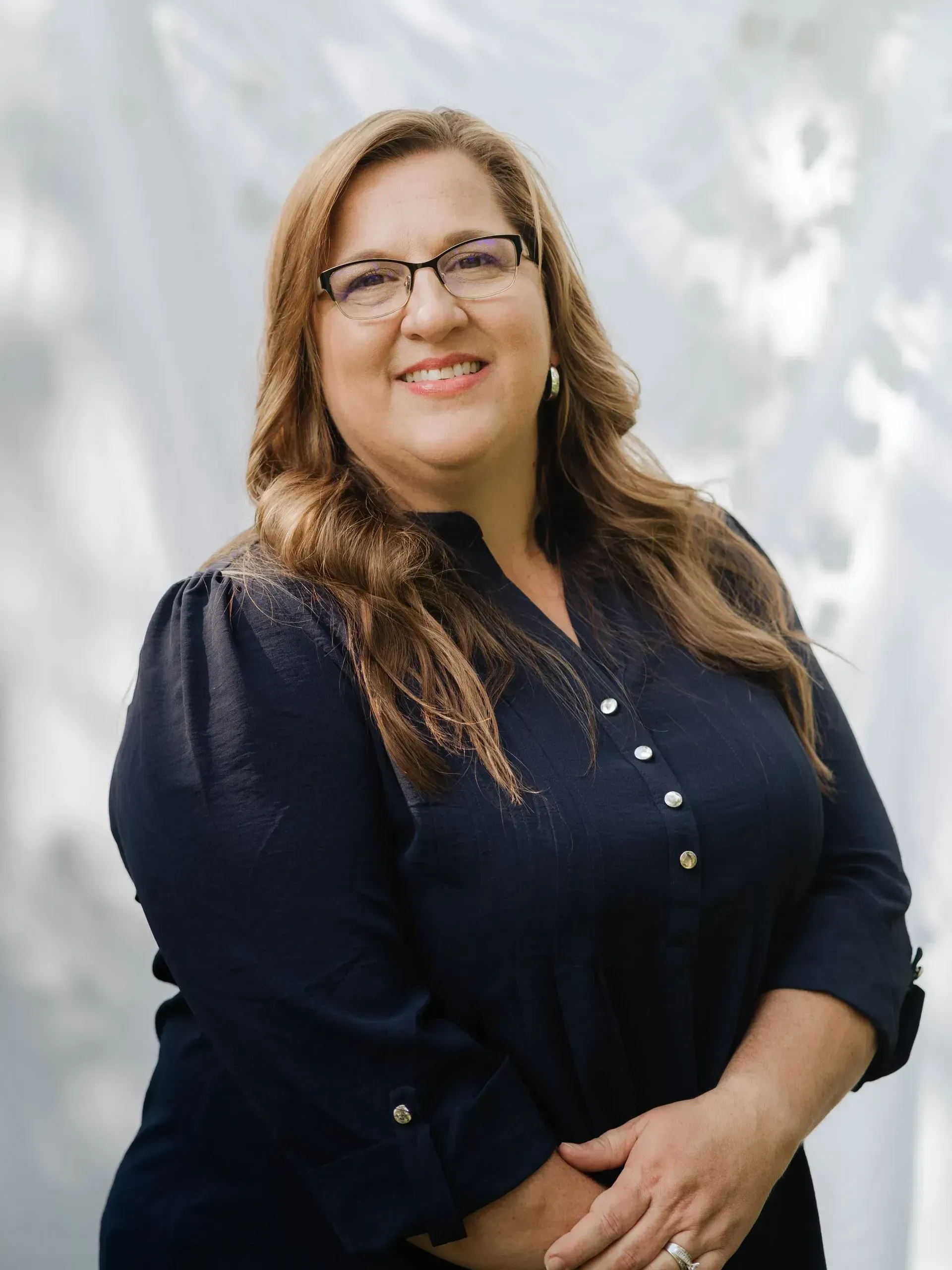 Woman with glasses smiles, wearing a navy blouse, outdoors.