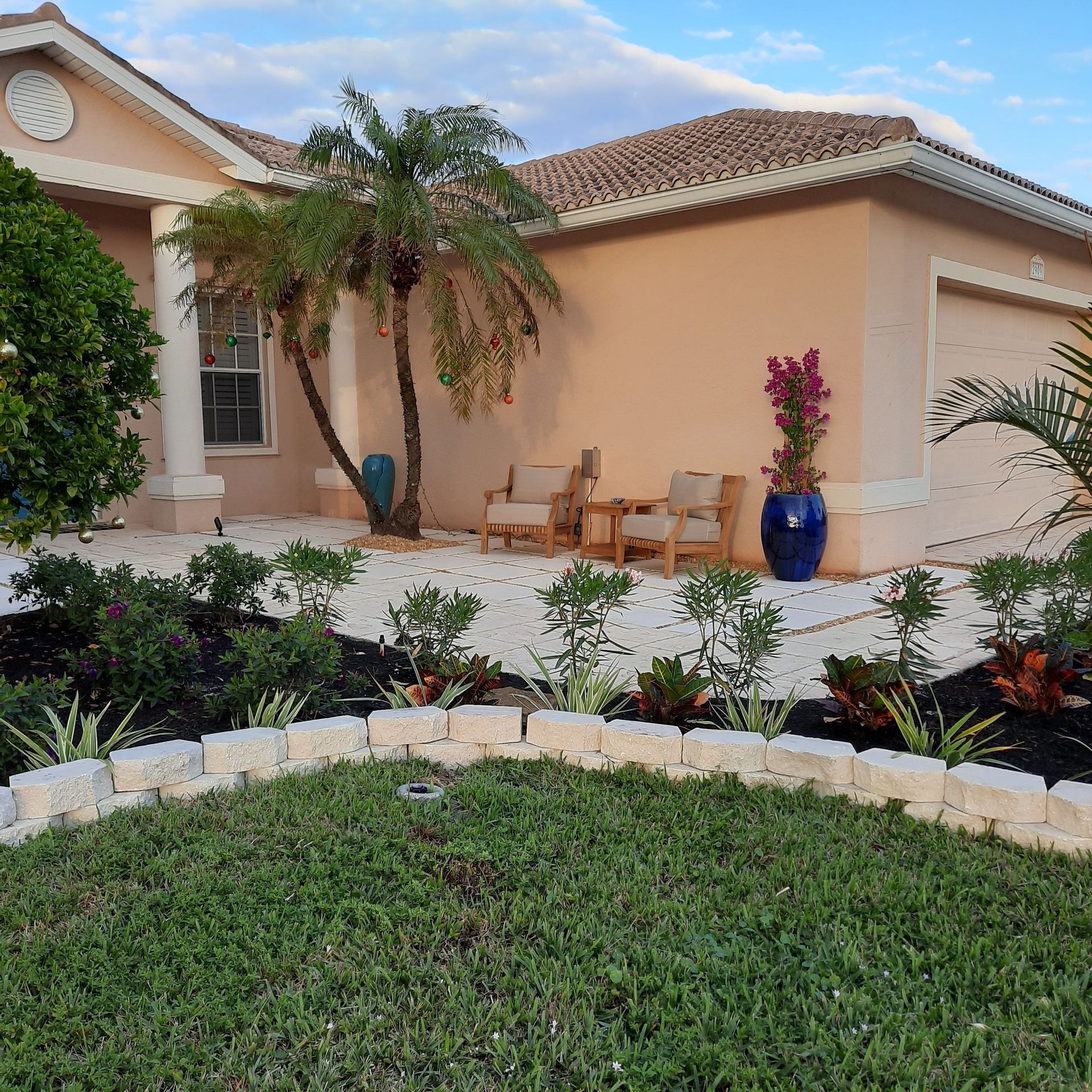 Front yard of a beige house with tiled roof, patio seating, palm tree, and colorful potted flowers.