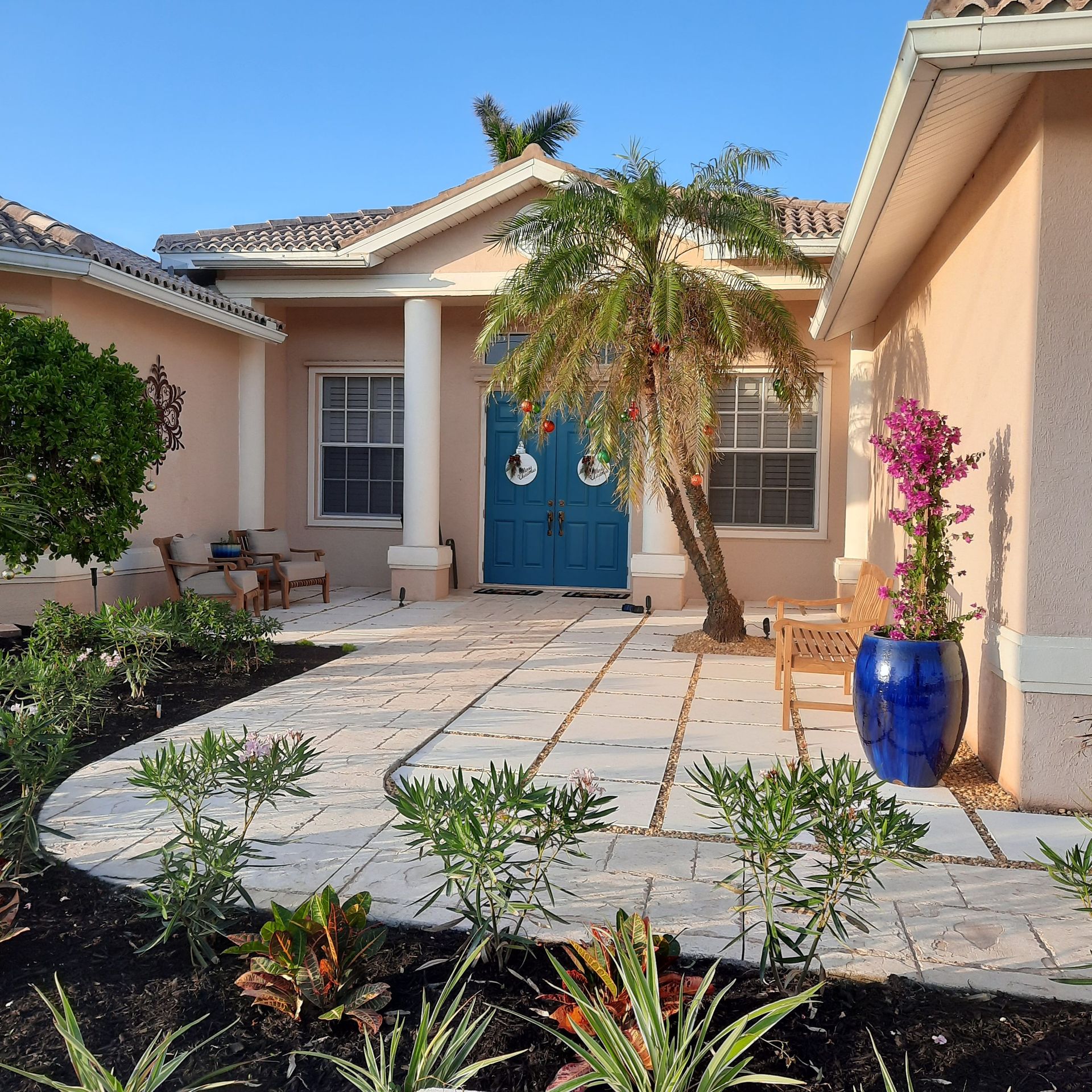 Front courtyard of a beige house with blue doors, tiled path, potted plants, and a flowering tree