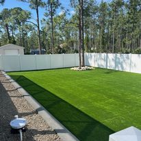 Lush green artificial turf lawn enclosed by a white fence. A shed and trees are in the background.