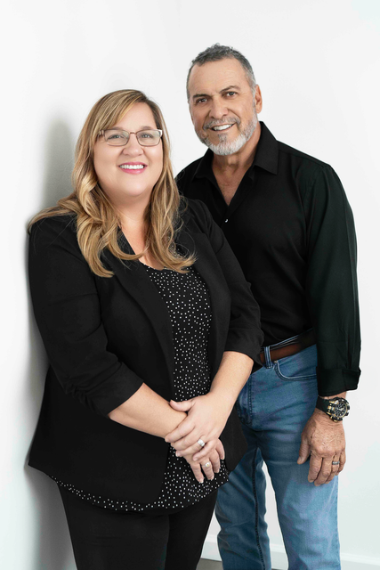 Woman and man posing for photo against white wall. Woman wears glasses, black jacket and dotted top; man wears black shirt and jeans.