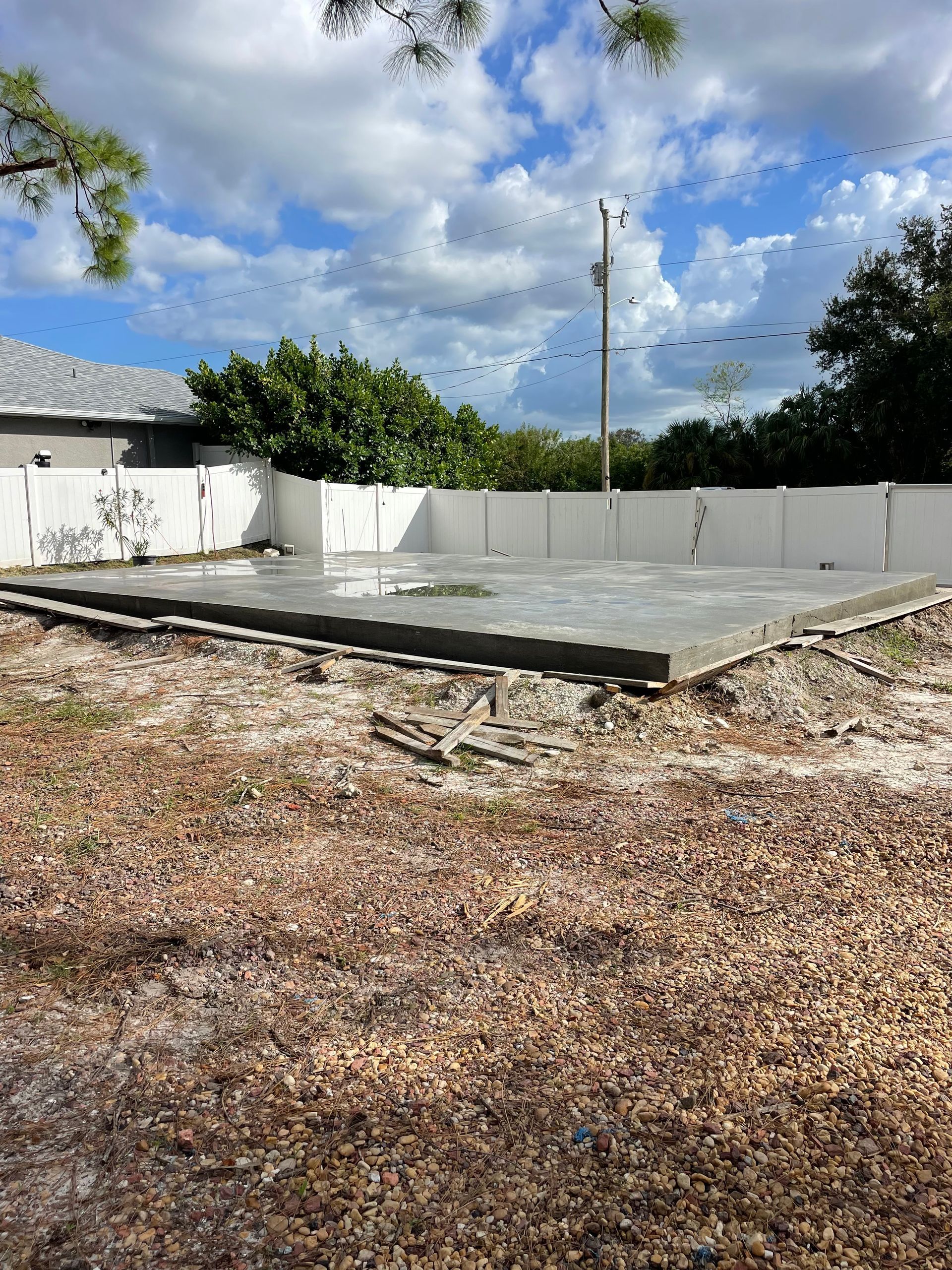 Concrete foundation surrounded by a white fence in a yard with brown dirt and vegetation under a cloudy blue sky.