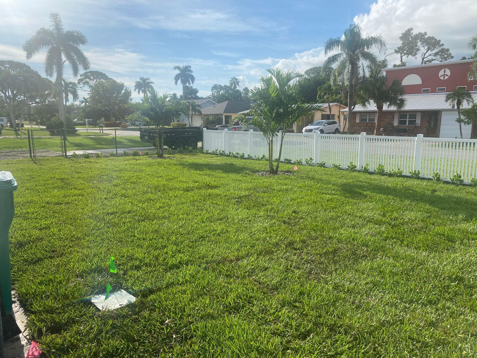 Lush green lawn with white picket fence, palm trees, and houses under a partly cloudy sky.