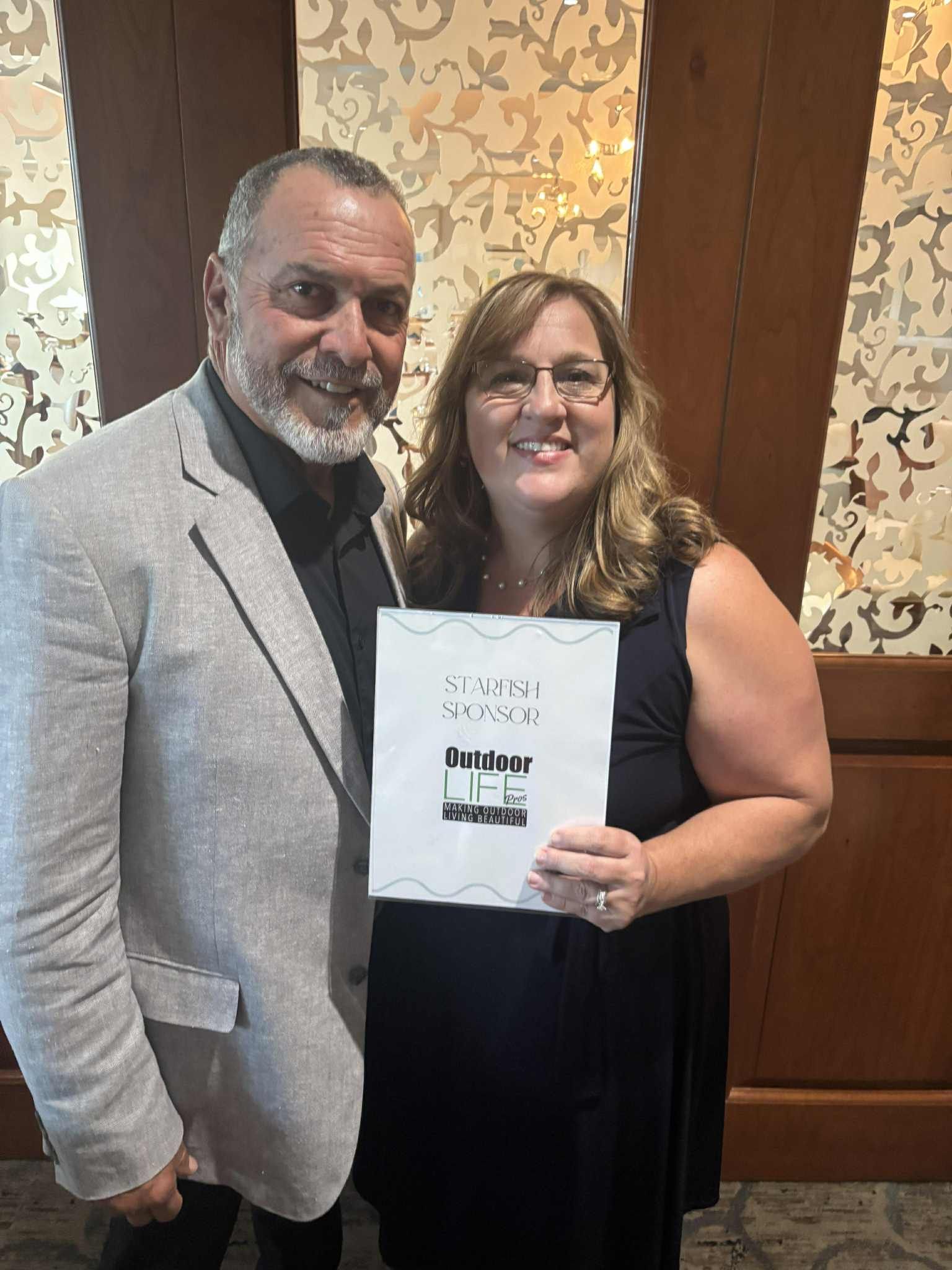 Man and woman smiling, holding certificate. They are in a formal setting with wood paneling.