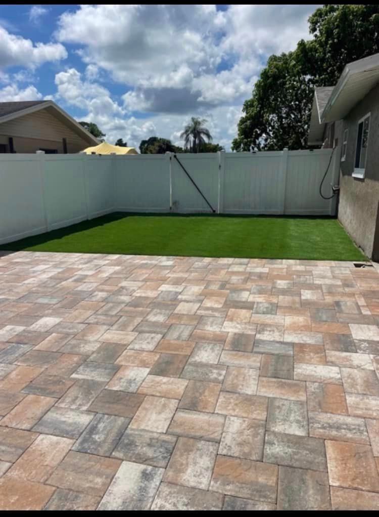 Patio with brick pavers, green lawn, white fence, and a house under a cloudy sky