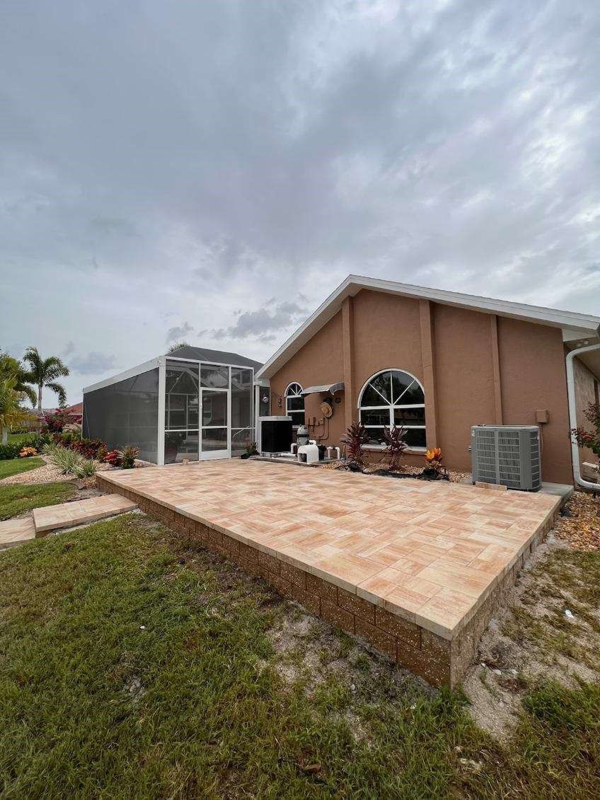 Tan patio and retaining wall beside a stucco house with a screened enclosure under a cloudy sky.