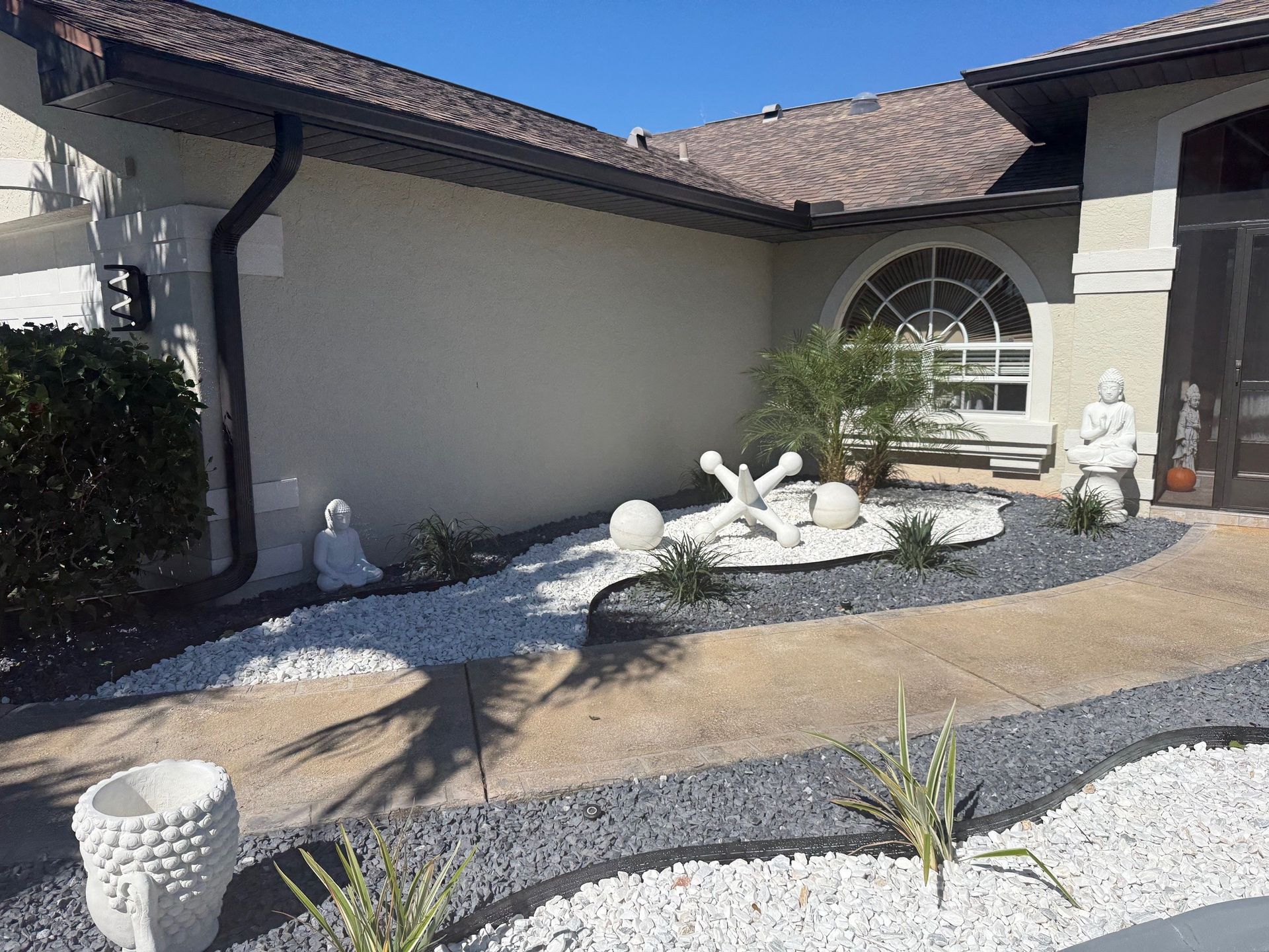 A modern landscaped front yard featuring white and gray rock beds, a large garden jack sculpture, and buddha statues.