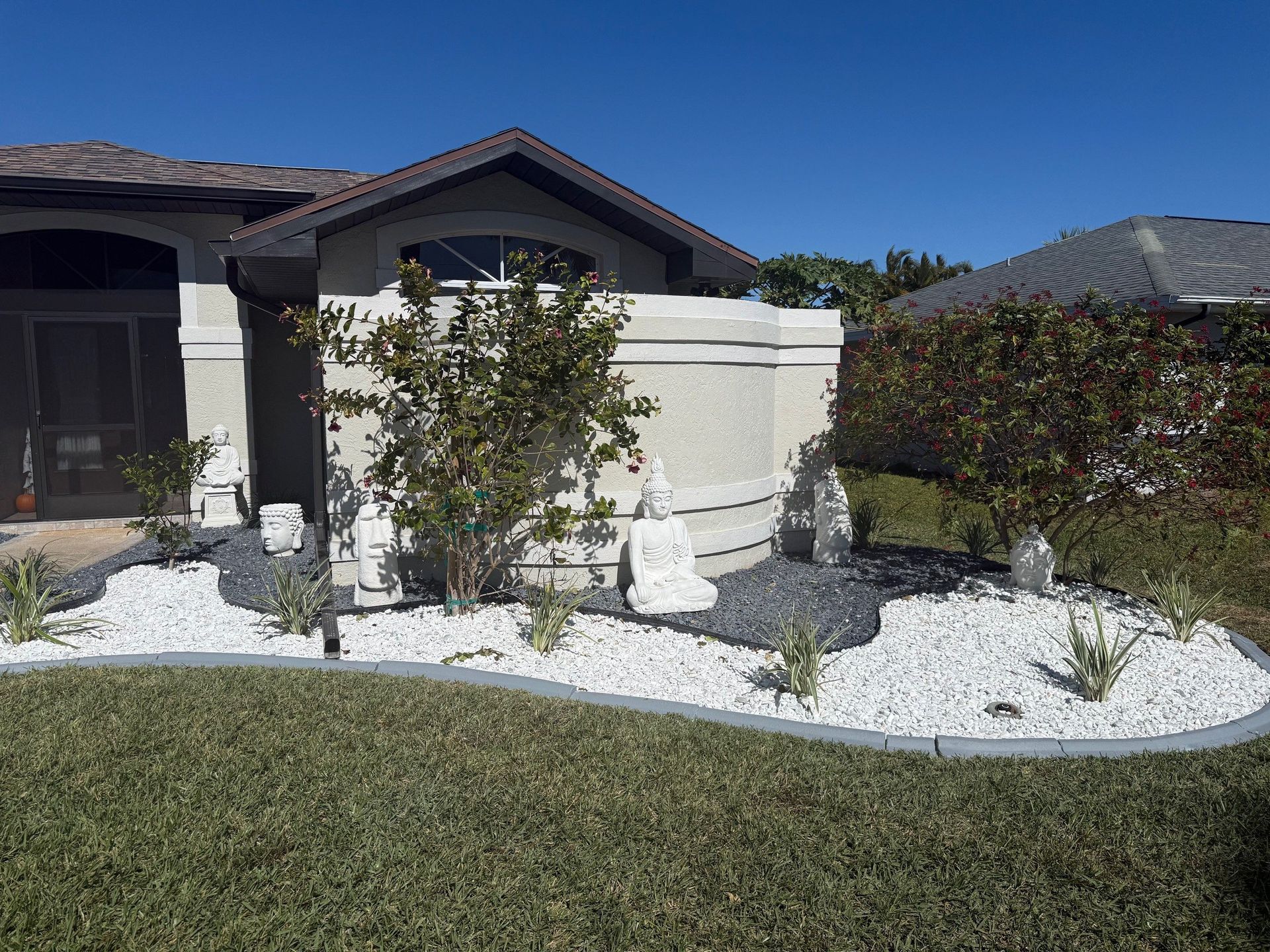 A curved landscaping bed in front of a house, featuring white stone, dark gravel accents, and several white Buddha statues.