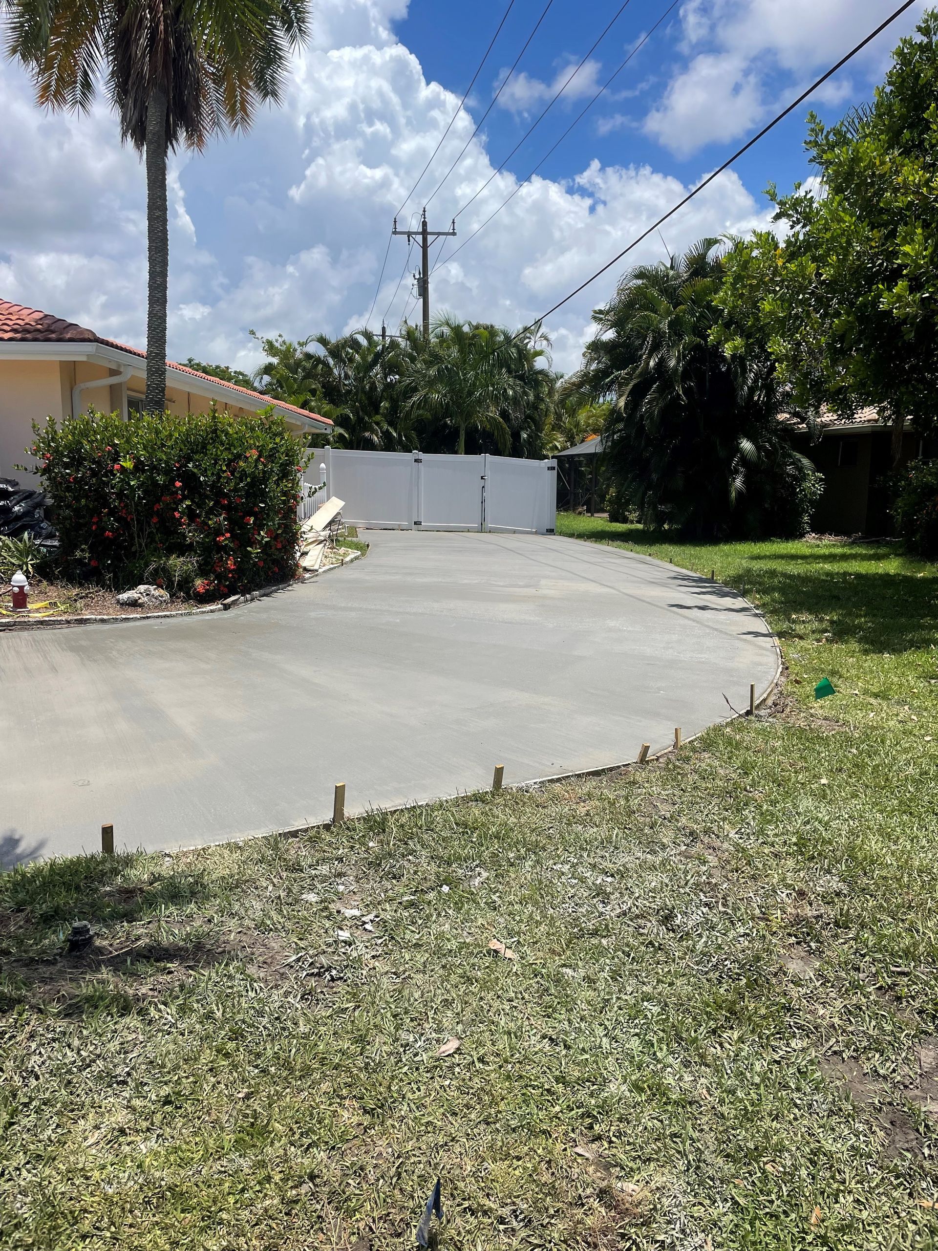 Asphalt driveway curves towards white gate, framed by lawn and trees under a blue sky.