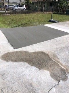 Gray concrete basketball court and driveway with water stain. Cars and houses in background.