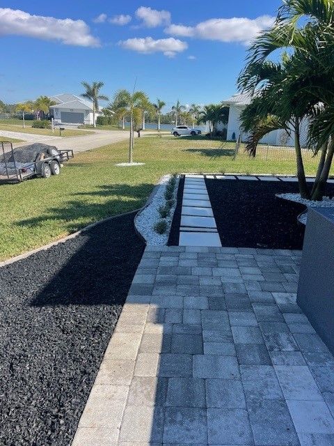 Path of gray pavers and stepping stones with dark mulch and white rock, in a yard with palm trees.