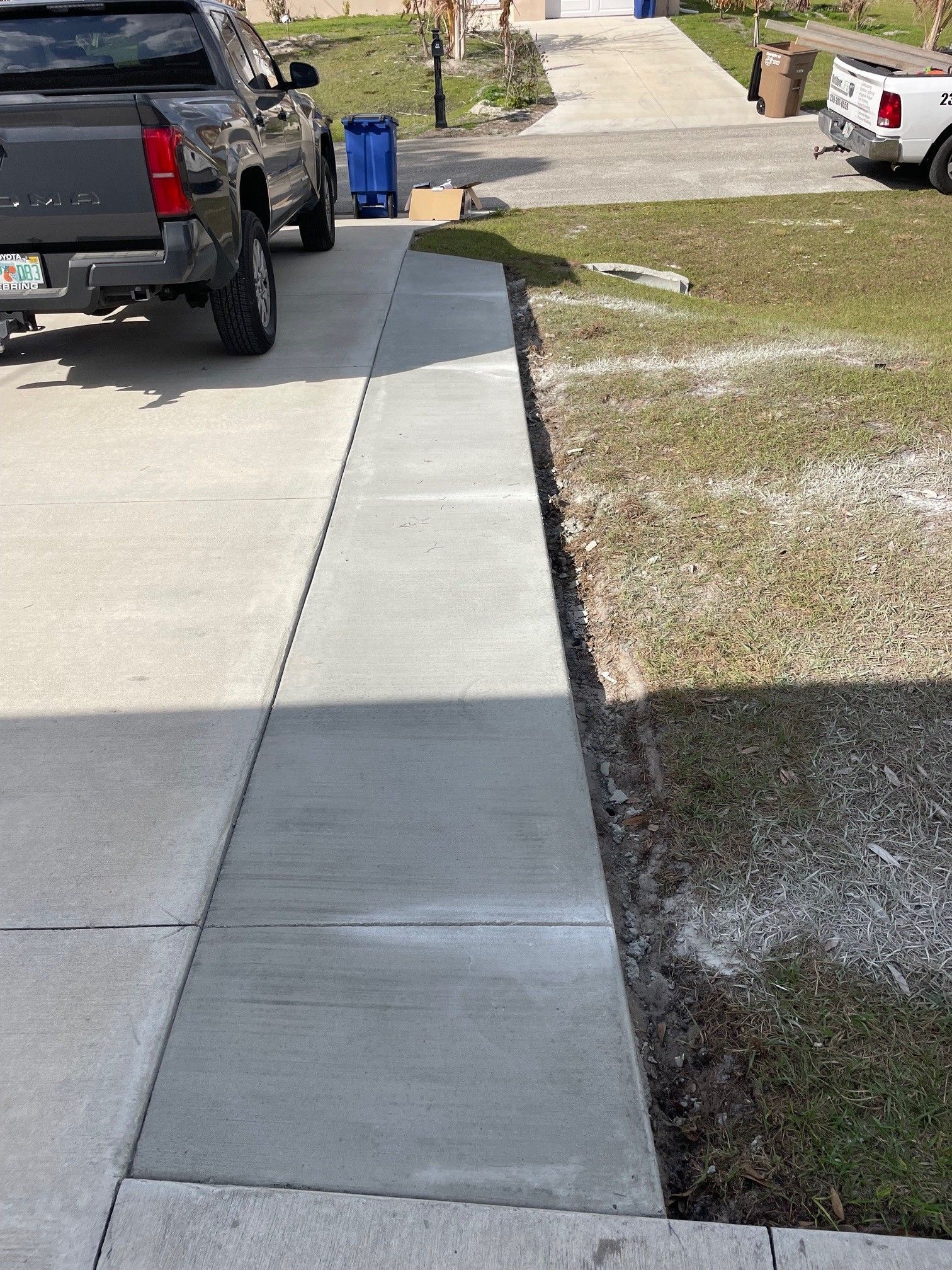 A newly poured concrete walkway extension runs parallel to a residential driveway, bordered by a strip of grass.