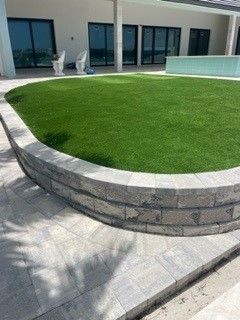 Curved gray brick wall surrounds a circular patch of green turf. Patio in foreground, large glass windows in background.