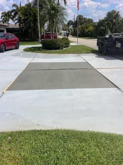 A freshly poured concrete driveway section on a sunny day with a red car and a trailer parked nearby.