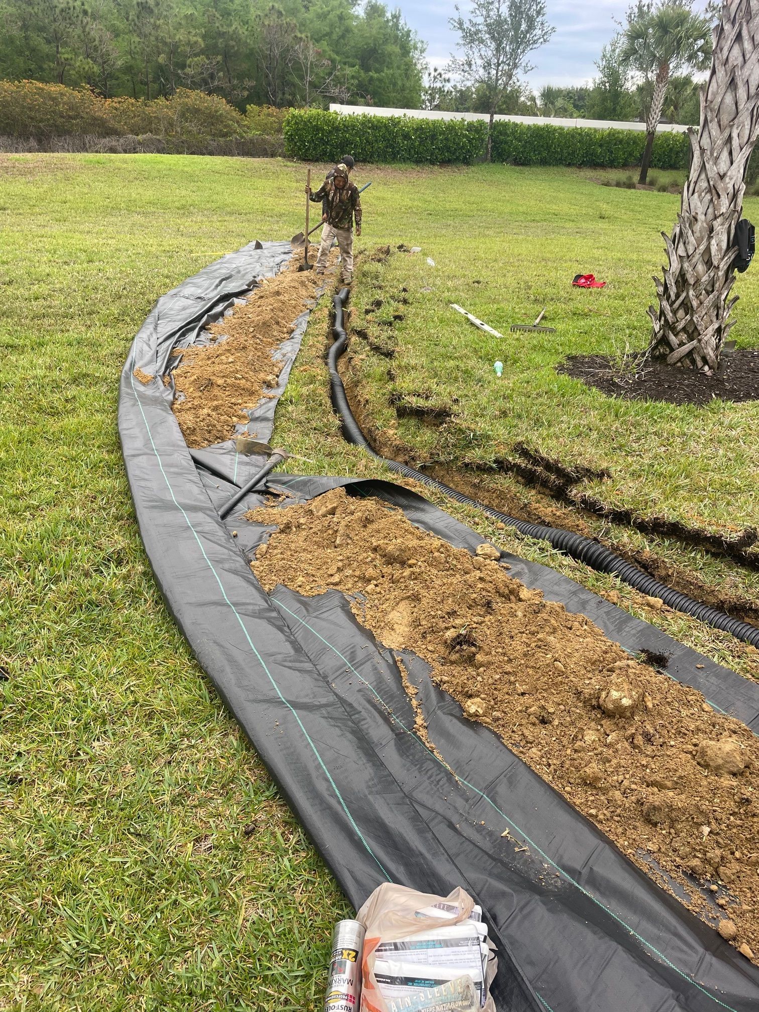 Man digging a trench, with black fabric and dirt laid out in a yard, near a tree.