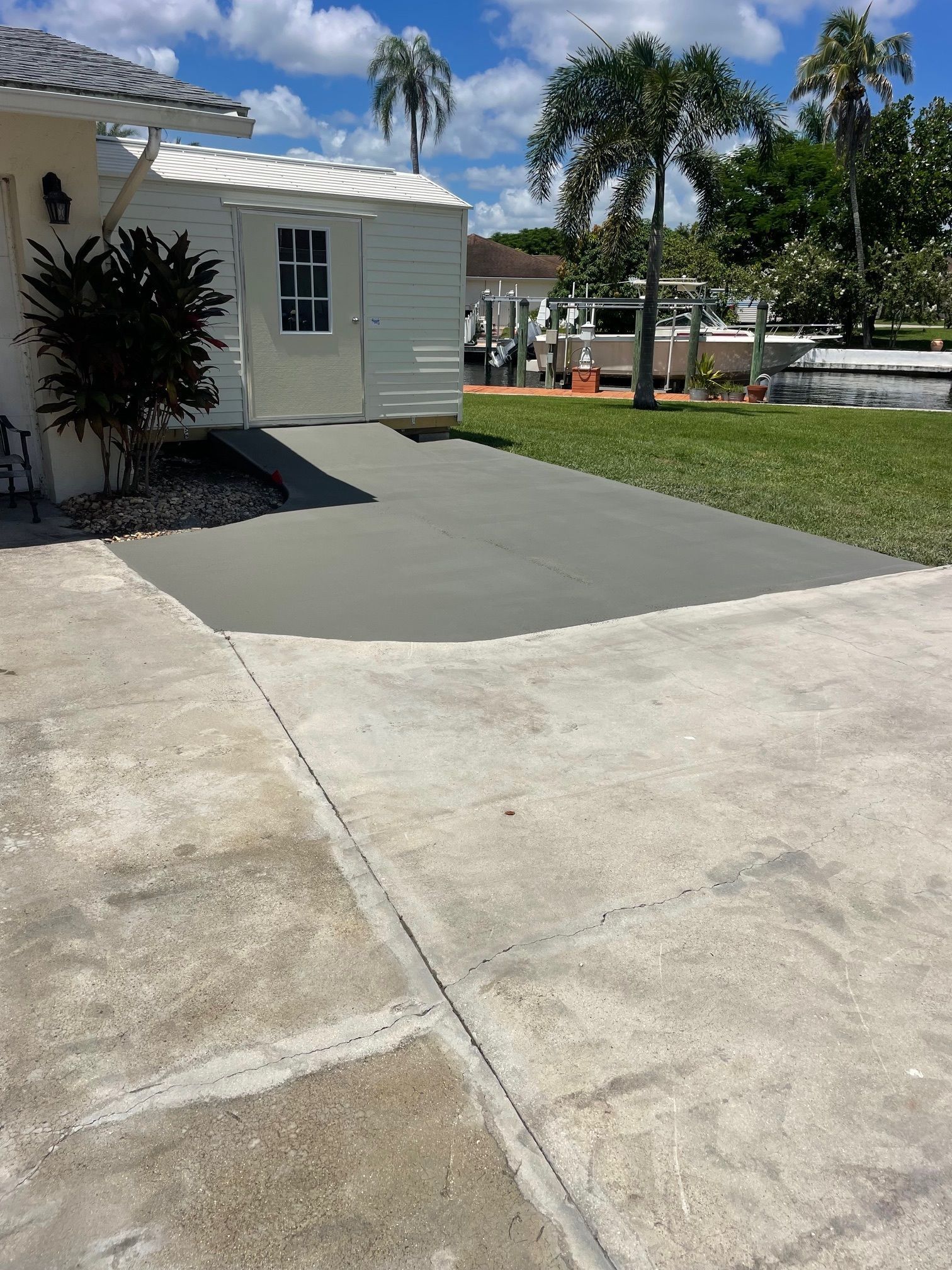 Driveway with a new gray concrete section next to a white building, blue sky and palm trees.