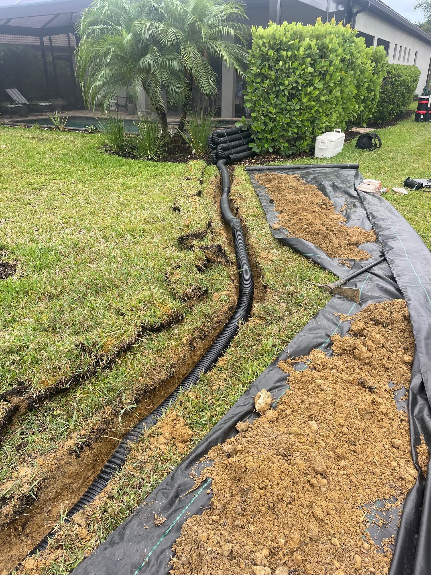 A trench with a black drain pipe being installed in a grassy yard. Brown soil is next to the trench.