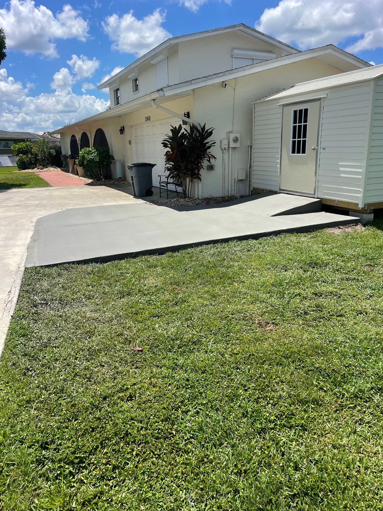 Side view of a cream-colored house with a shed and driveway; green grass and a blue sky.