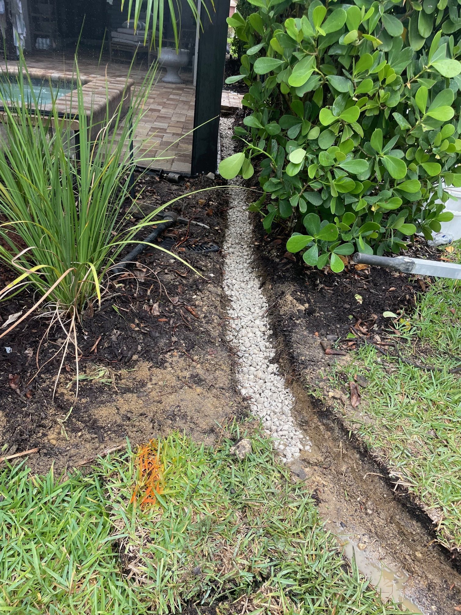 Gravel-filled trench along a yard's edge, near greenery and a dark post.