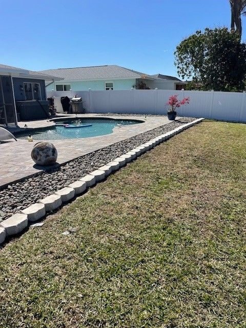 Backyard with pool, grass, rocks, and white fence on a sunny day.