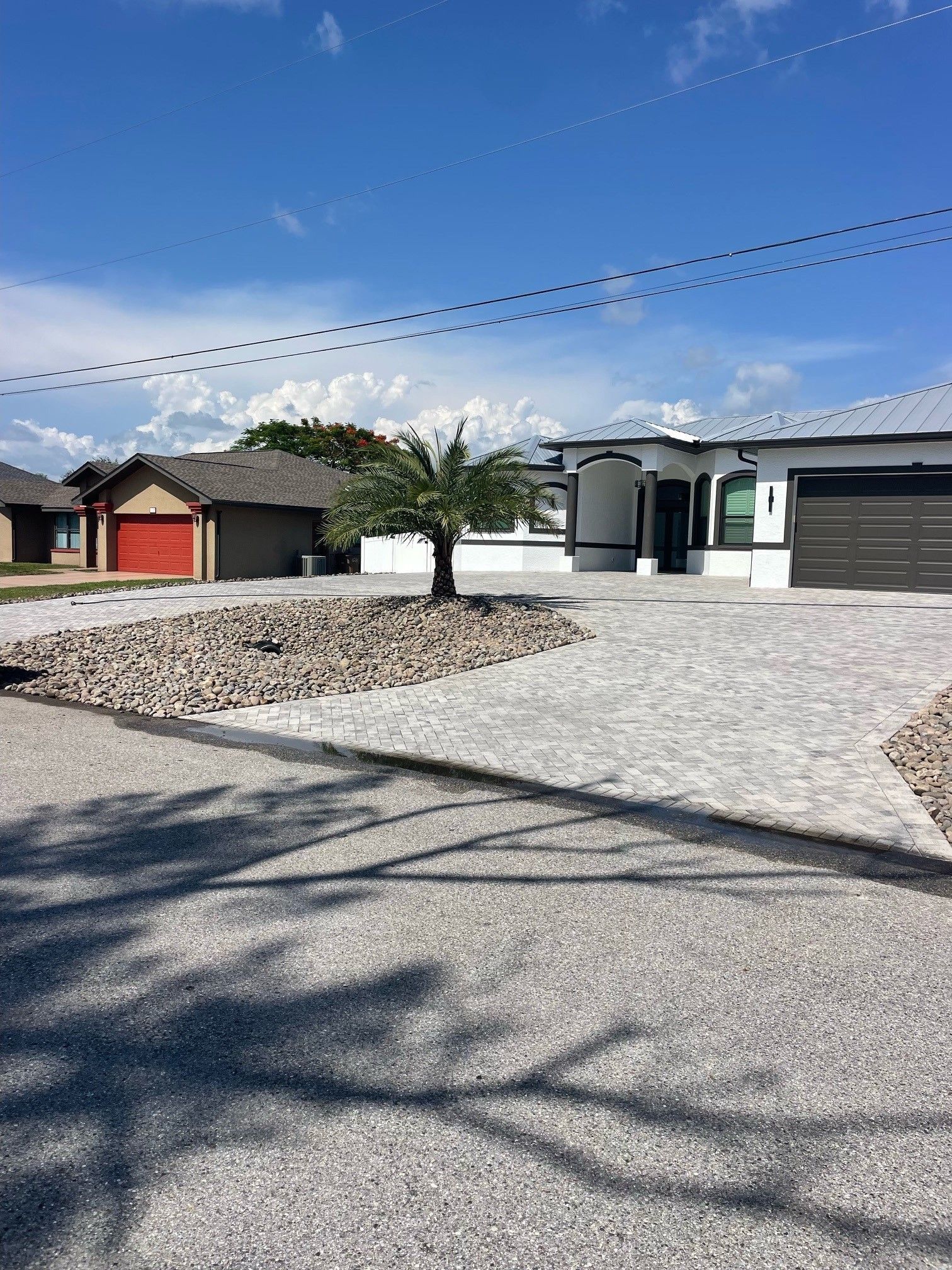 A modern house with a gray driveway, a small palm tree, and a red garage in the distance.