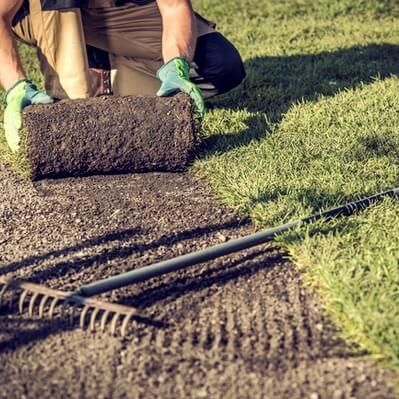 Person kneeling, rolling sod on prepared ground. Green gloves and grass, brown soil, rake present.