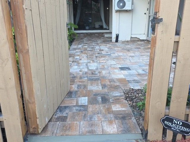 A stone paver patio viewed through an open wooden gate toward the entrance of a house.