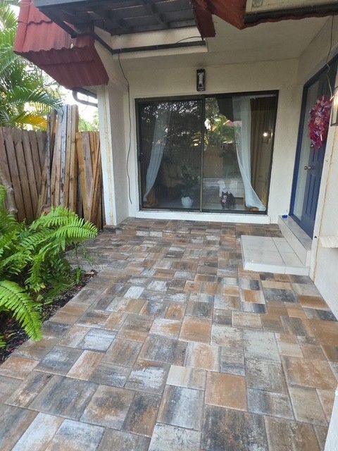 A stone-paved patio area leading to glass sliding doors of a home, flanked by a lush green plant and a wooden fence.