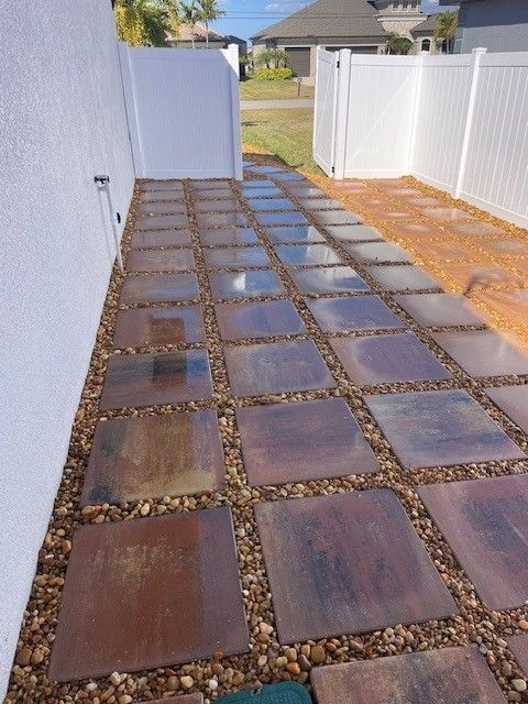A pathway of square stepping stones separated by gravel, located in a side yard between a house wall and a white fence.