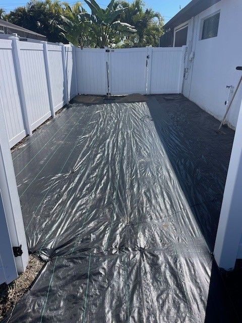 A black landscape fabric covers a rectangular ground area between a white vinyl fence and a house wall.