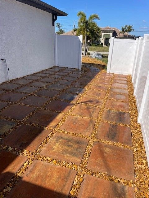 A side yard with a patio of square stone pavers separated by gravel strips, running between a white house wall and fence.