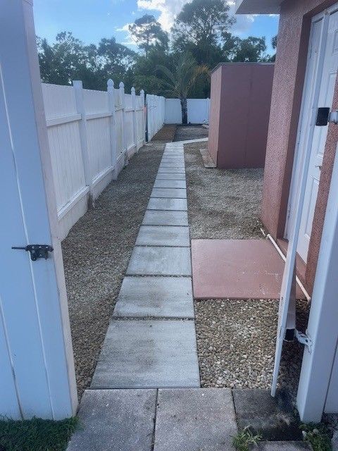 A narrow pathway made of concrete slabs leads through a yard lined with a white fence and gravel.