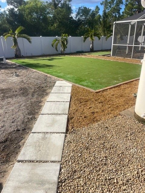 Backyard landscaping with artificial turf, stepping stones, and gravel. Palm trees and a screened enclosure are visible.