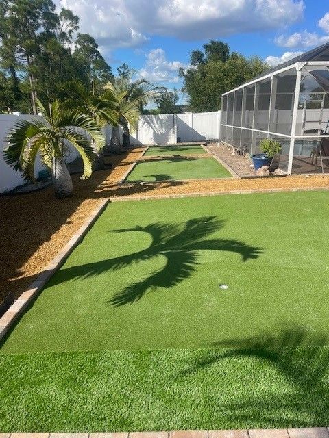 Artificial turf putting green in backyard with palm tree shadow. Fenced yard, sunny day.