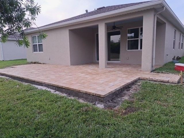 Backyard patio with brick pavers, tan walls, and a covered porch. Green grass surrounds.