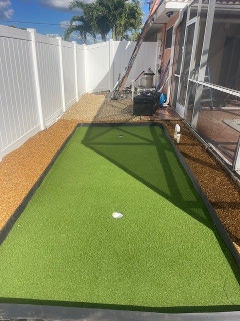 A backyard putting green with a white fence on one side and a home on the other, sunny day.