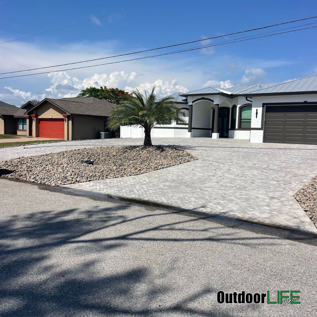 House with paved driveway and rock landscaping. Palm tree in the center. Clear sky.