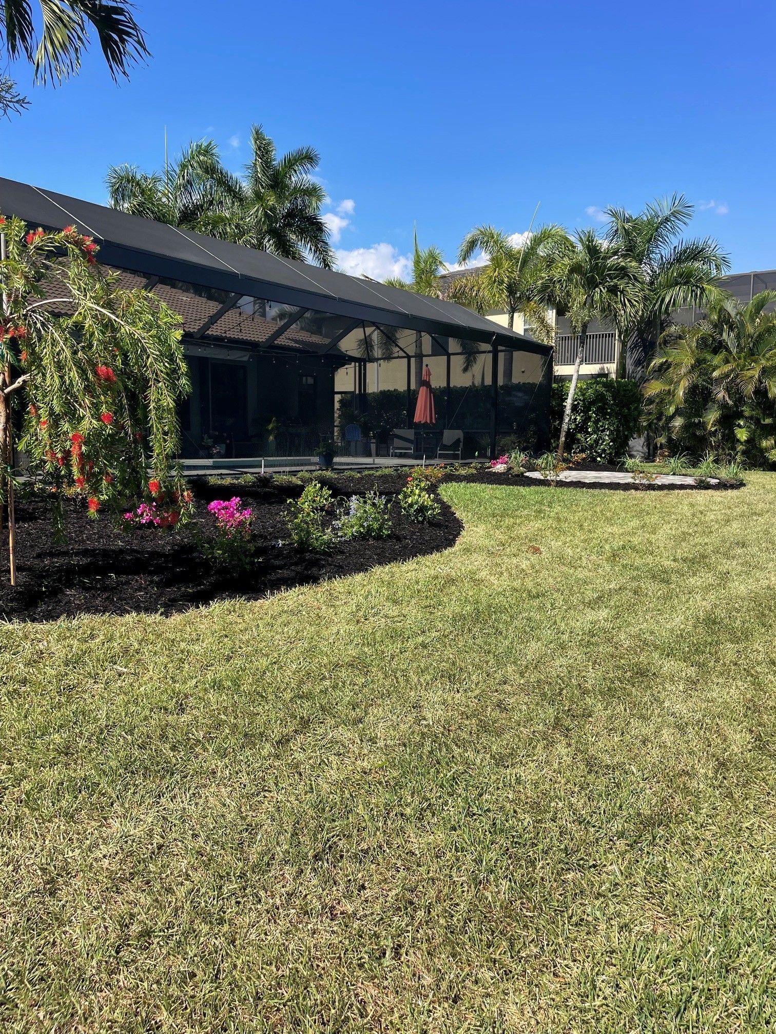 Single-story house with screened porch, palm trees, and a sunny front lawn with flower beds.