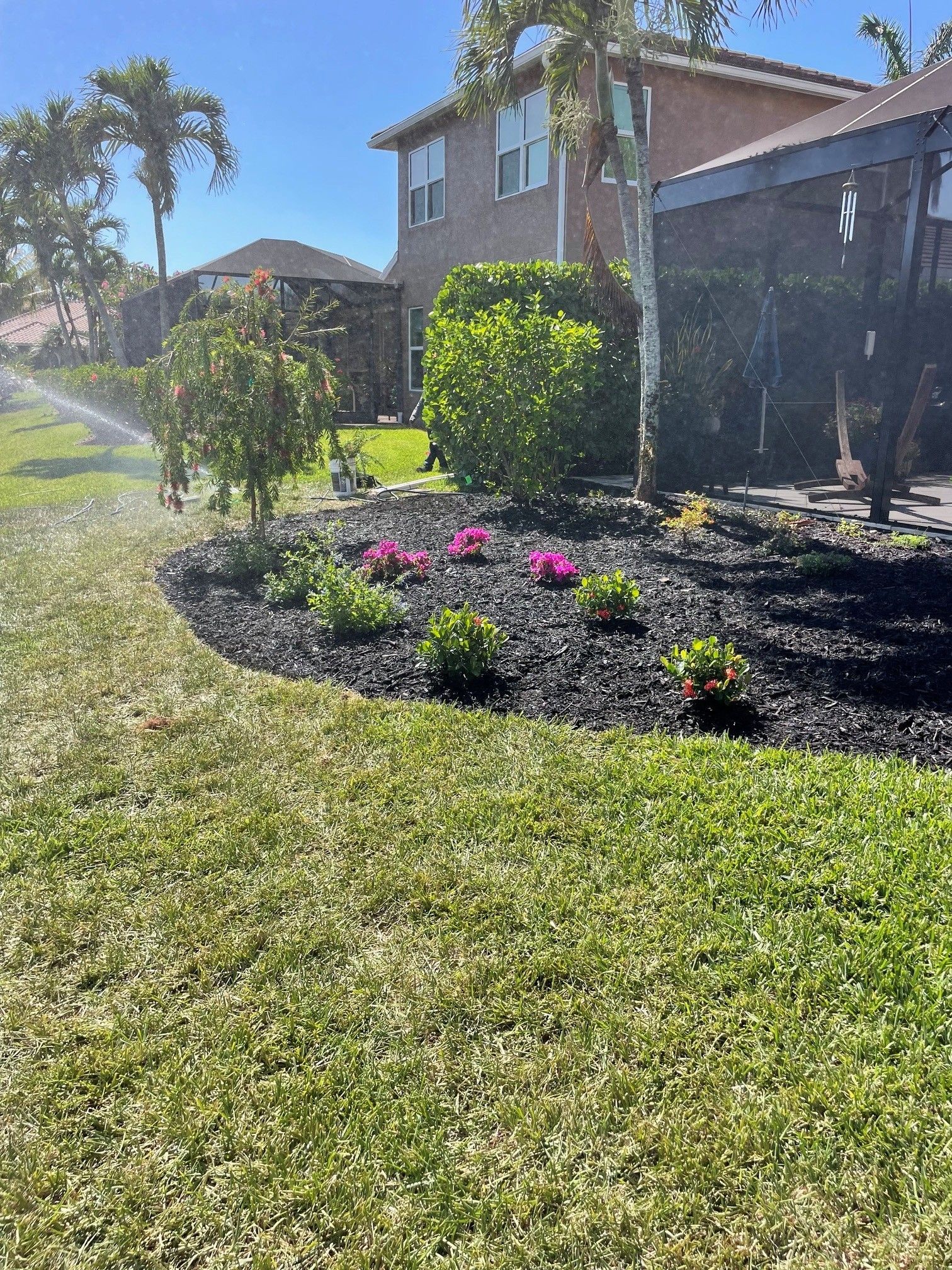 Sunny suburban yard with a flower bed of pink blooms and trimmed shrubs beside a brick house