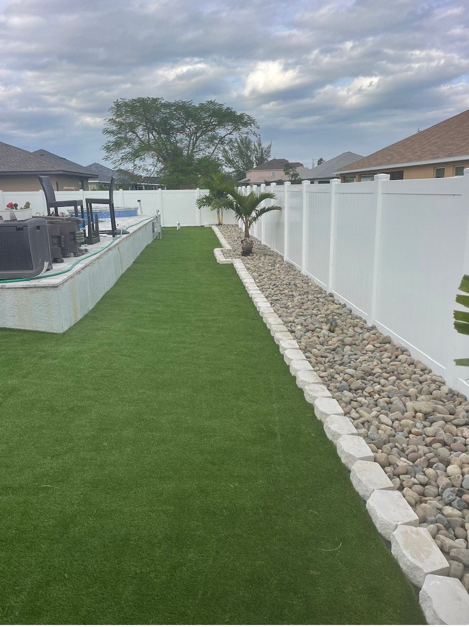 Backyard with green turf, white fence, rock border, and cloudy sky.