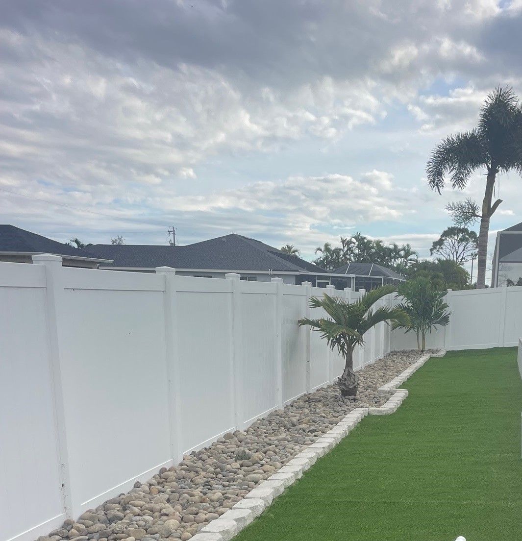 White vinyl fence surrounds a backyard with a grassy lawn, rock border, and a palm tree against a cloudy sky.